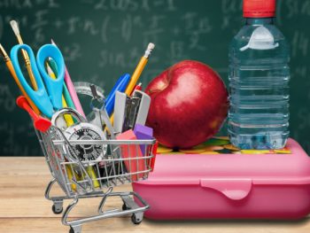 A shopping cart filled with school supplies next to an apple and water bottle on top of a lunch box.